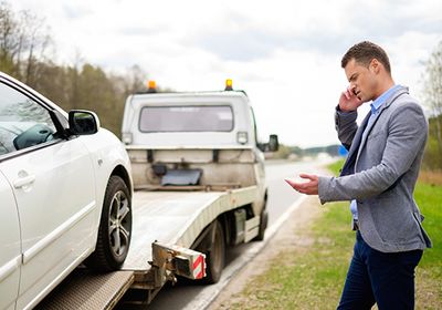 Geschäftsmann steht telefonierend am Fahrbahnrand im Hintergrund wird sein Auto abgeschleppt