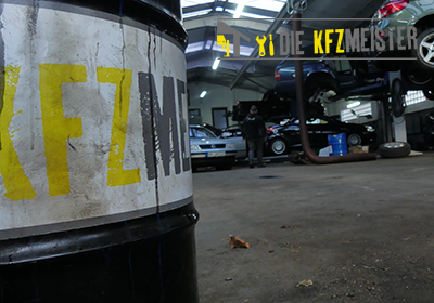 Vordergrund: Ölfass mit KFZ Meister Schrift Hintergrund: KFZ-Meisterwerkstatt